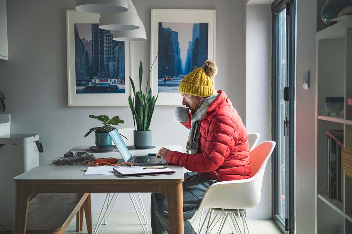 Mature man sitting working from home in a red puffer coat, scarf and wooly hat - Cost of Living Crisis